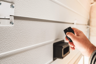 Close-up of a hand pointing a garage door remote control at a wall-mounted sensor unit